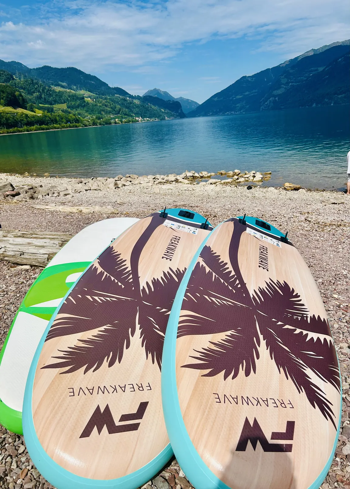 Mietboards liegen bereit am Kiesstrand, dahinter der türkisblaue See und die Berge